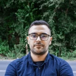 Professional portrait of a man wearing glasses and a dark shirt, standing outdoors with arms crossed, looking confidently to the side.