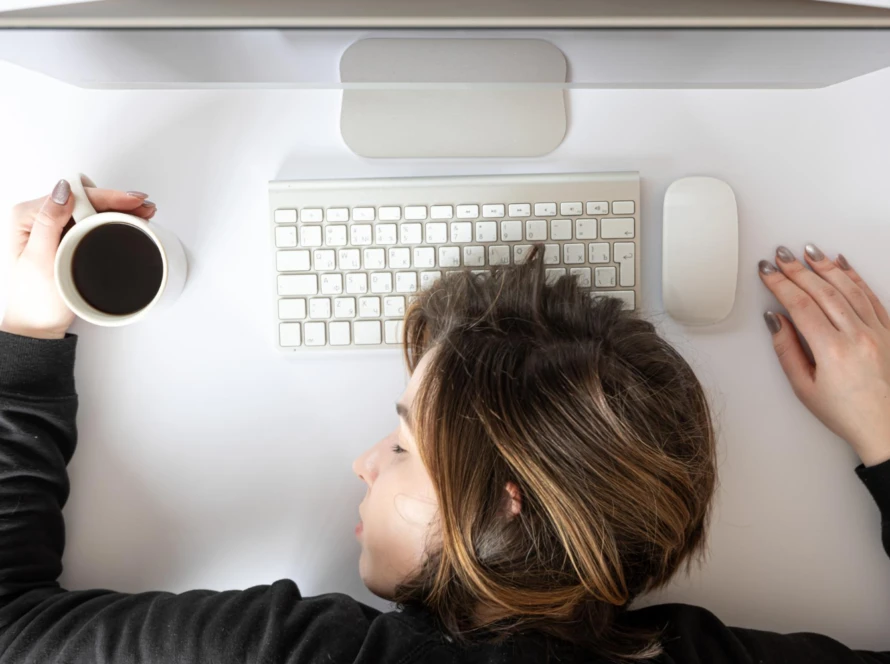 Exhausted software developer asleep on a white desk, holding a cup of black coffee beside a computer keyboard and mouse, illustrating burnout from sprint-crunch overtime.