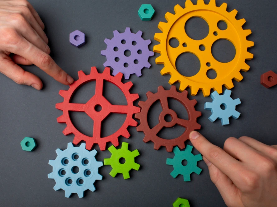Colorful wooden gears of different sizes being aligned by two hands on a dark surface—visual metaphor for how a Scrum Master meshes people, process and just-enough technical insight.