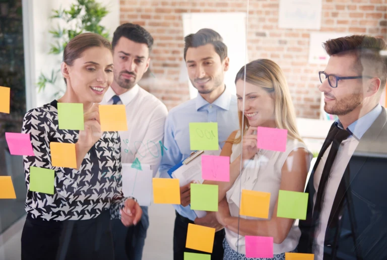Team of five reviewing a glass board covered with colorful sticky notes, discussing WIP limits and next steps.