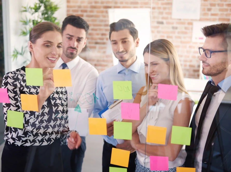Team of five reviewing a glass board covered with colorful sticky notes, discussing WIP limits and next steps.