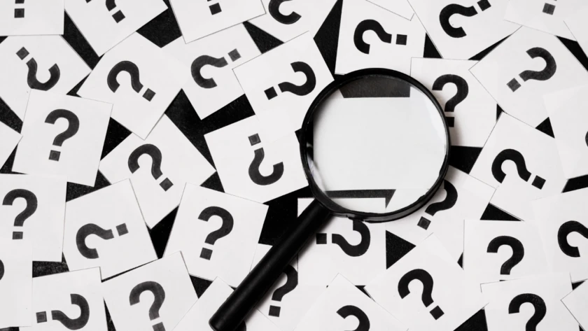 Magnifying glass lying on scattered white cards printed with bold black question marks.