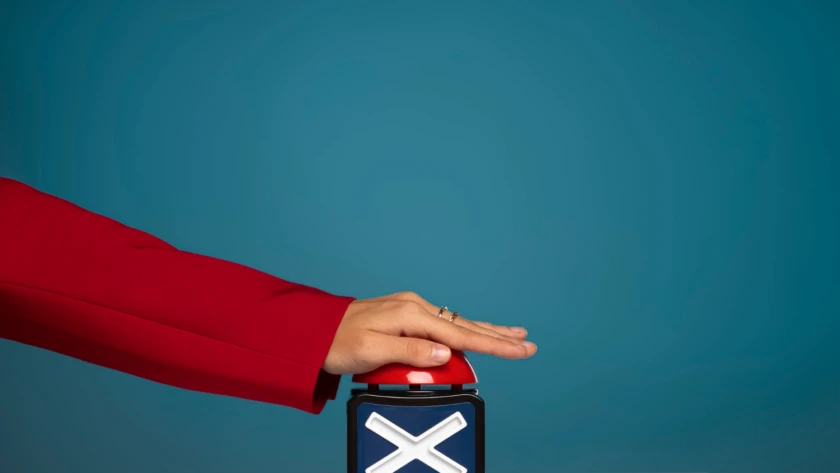 Hand in a red blazer pressing a large red buzzer on a box with a white “X,” against a teal background.