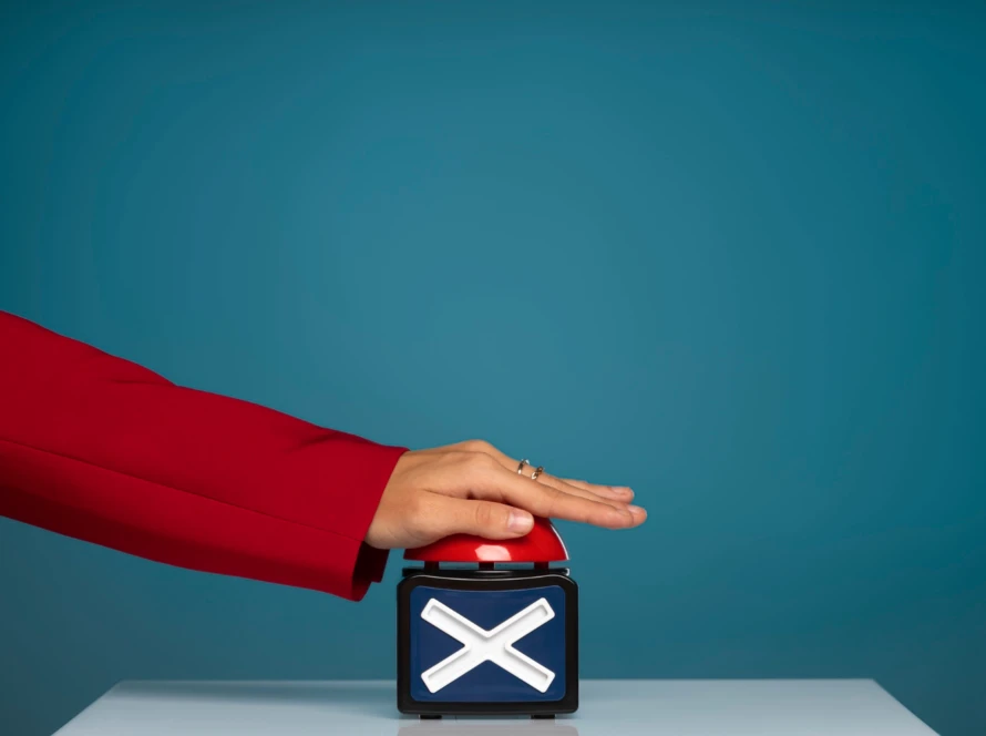Hand in a red blazer pressing a large red buzzer on a box with a white “X,” against a teal background.