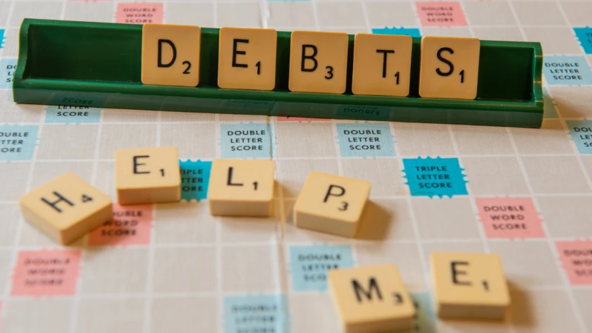Scrabble board with tiles—rack reads “DEBTS” and loose tiles spell “HELP ME.”